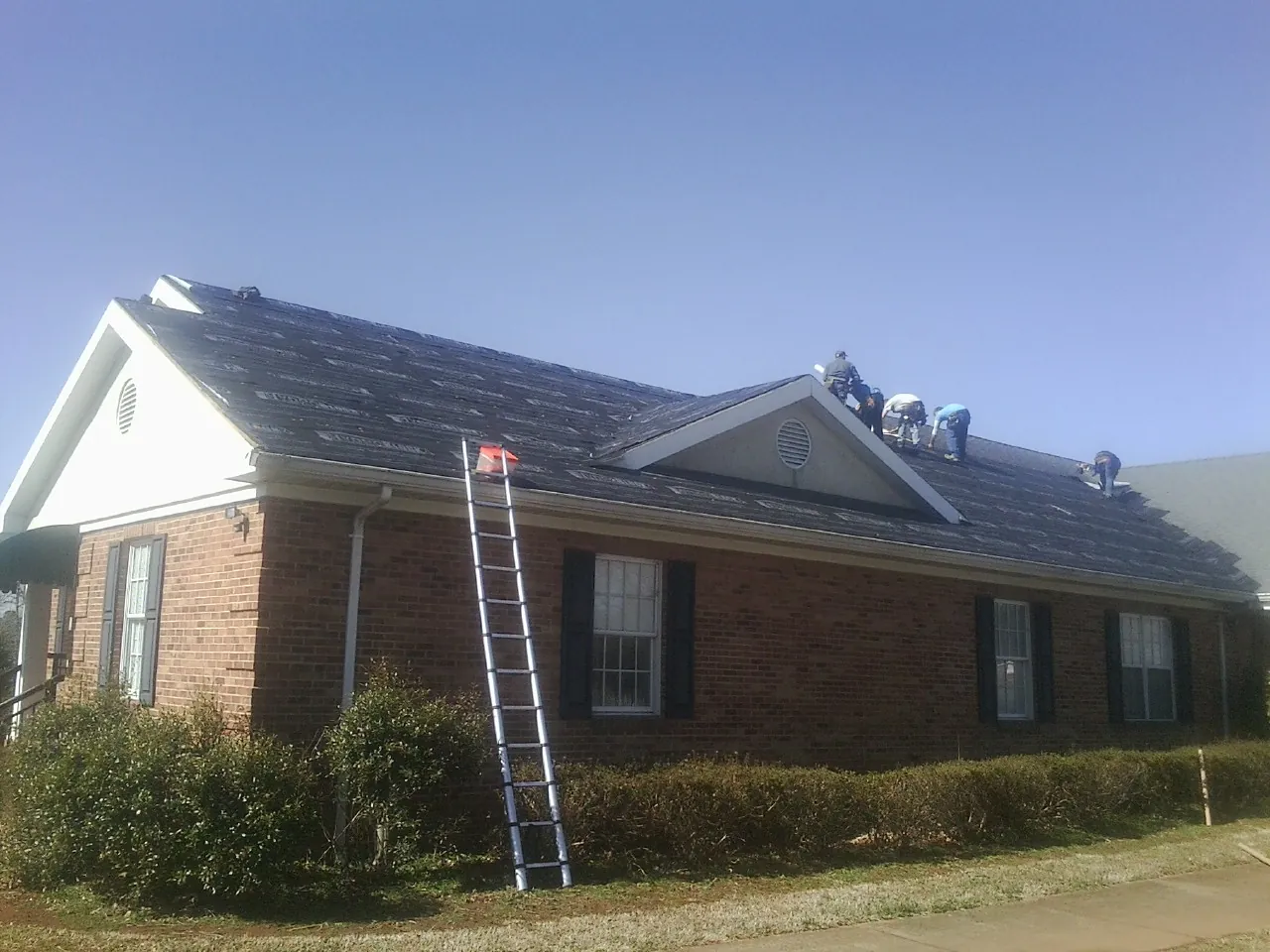 Crew tearing shingles off a brick ranch roof with a ladder set — Emergency Roof Repair work by our crew in Manchester
